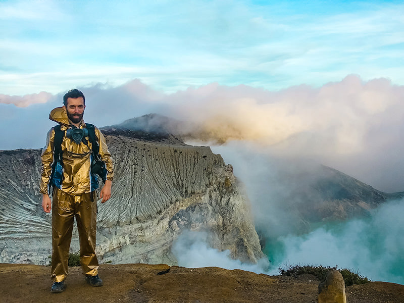 Pipeaway blogger Ivan Kralj dressed in golden gear standing on the edge of the Ijen Volcano in East Java, clouds, mountain and blue crater lake visible in the background
