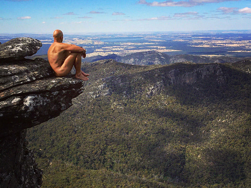 Erik, the Australian naked hiker, sitting on the edge of the cliff, with a beautiful mountain forest view