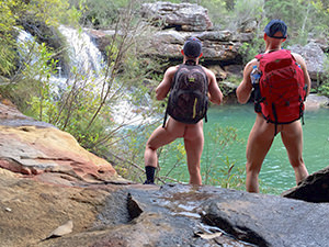 Photograph of two naked hikers with backpacks in front of the waterfall