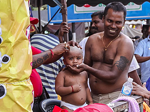 Photograph of a father holding his toddler son's head while a barber is shaving him bald for Thaipusam festival, photo by Ivan Kralj