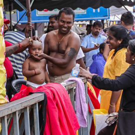 Photograph of a street barber shaving bald one of the Thaipusam youngest pilgrims - toddler's head is being held still by his father, while other relatives watch and laugh, photo by Ivan Kralj