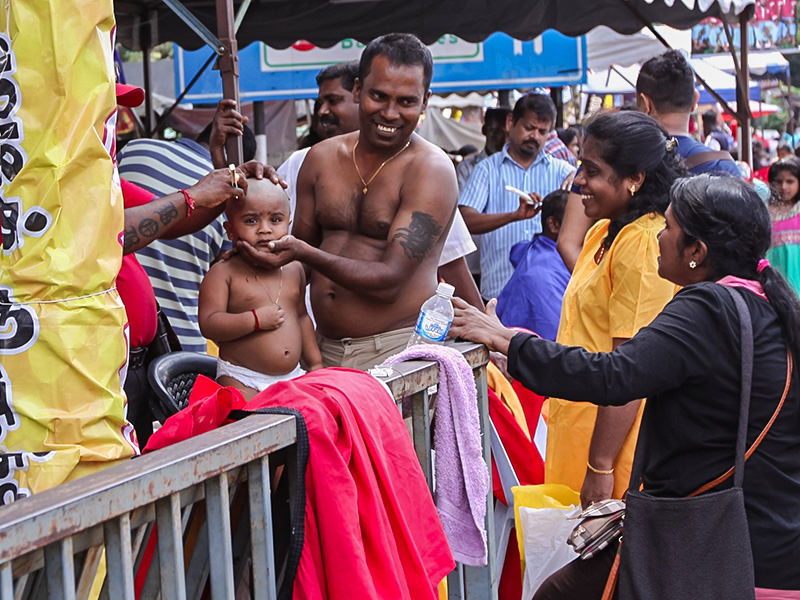 Photograph of a street barber shaving bald one of the Thaipusam youngest pilgrims - toddler's head is being held still by his father, while other relatives watch and laugh, photo by Ivan Kralj