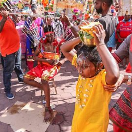 Photograph of a girl carrying a Paal Kudam, a pot of milk, spilling over her forehead, while her mother supports her at Thaipusam pilgrimage. At the background of the photo, a young guy is resting on chair while carrying a Kavadi, a heavy portable altar with skewers attached to his torso, photo by Ivan Kralj