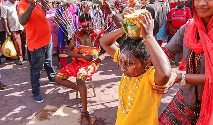 Photograph of a girl carrying a Paal Kudam, a pot of milk, spilling over her forehead, while her mother supports her at Thaipusam pilgrimage. At the background of the photo, a young guy is resting on chair while carrying a Kavadi, a heavy portable altar with skewers attached to his torso, photo by Ivan Kralj