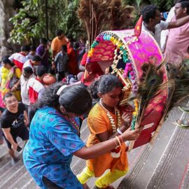 Photograph of a mother helping her young daughter carrying a heavy Kavadi altar while climbing the stairs of Batu Caves for Thaipusam festival, photo by Ivan Kralj