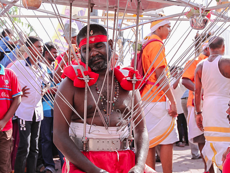 Photograph of a Thaipusam pilgrim resting while wearing a Kavadi, a portable altar at Thaipusam - the structure is worn on his shoulders while the skewers are pressing the bearer's torso, photo by Ivan Kralj
