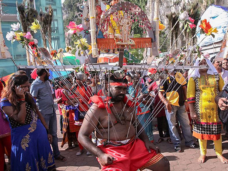 Photograph of a Thaipusam pilgrim resting while carrying a Kavadi, a portable altar at Thaipusam - the structure is worn on his shoulders while the skewers are pressing the bearer's torso, photo by Ivan Kralj