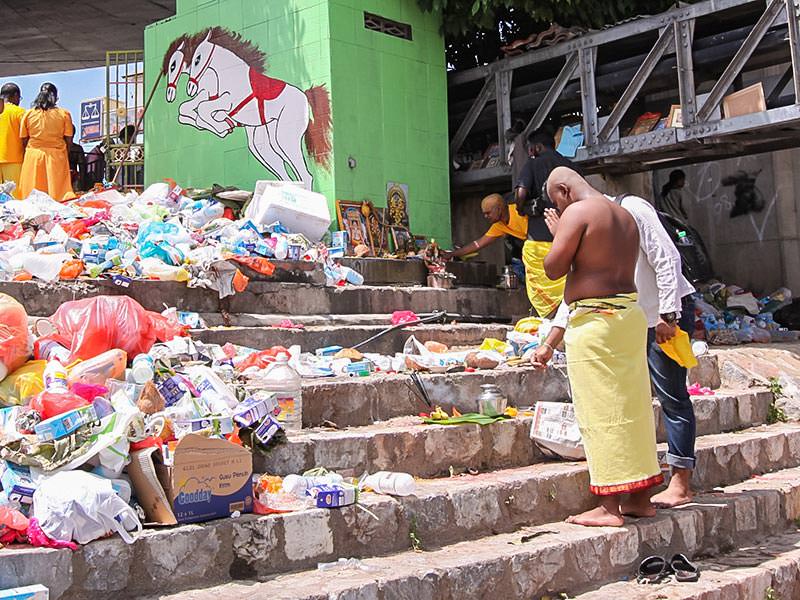 Photograph of a pilgrim praying in front of the improvised altars in piles of trash at the river bank at Thaipusam festival
