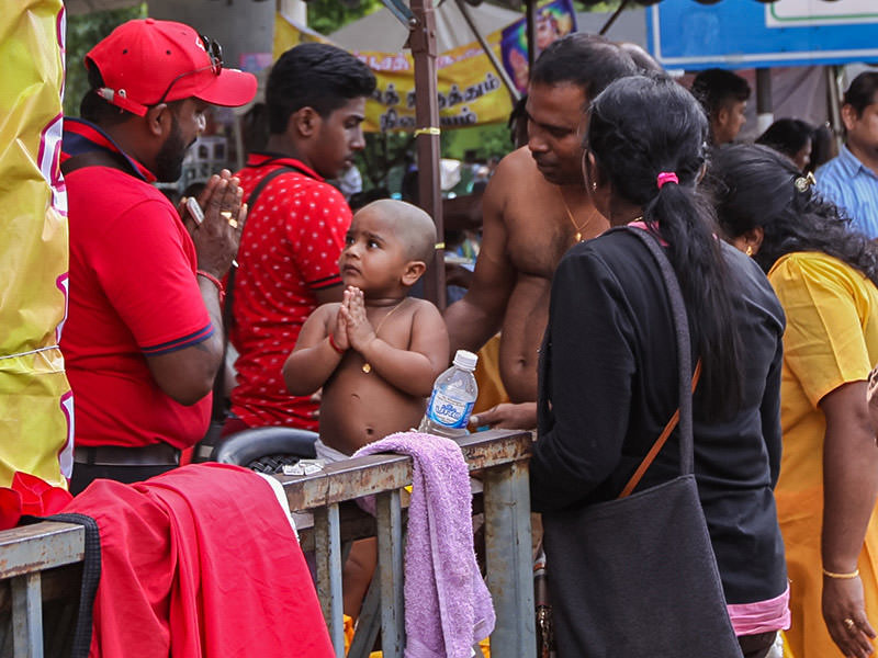 Photograph of a toddler giving a gesture of thanks to the street barber that shaved him bald at Thaipusam, photo by Ivan Kralj