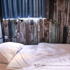 Photo of the bed in the dormitory of The Laban hotel, white bedsheets in front of the blue-grey planks covering the wall, photo by Ivan Kralj