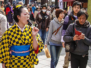 Asian lady in yellow-and-black checkered kimono licking a penis-shaped lollipop on the street of Kawaski, Japan, during Kanamara Matsuri festival, photo by Ivan Kralj.