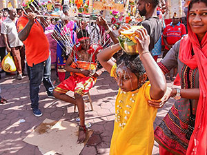 A little girl carrying milk-filled paal kudam on her head with mother supporting her, milk spilling on her forehead, while a young man, with spikes of kavadi piercing his skin, is resting in the background, at Thaipusam Festival in Batu Caves, Malaysia, photo by Ivan Kralj