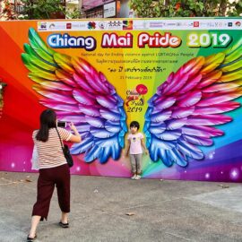 A mother taking a photograph of her child standing in front of the image of rainbow-colored wings at Chiang Mai Pride, gay parade in Chiang Mai, Thailand, photo by Ivan Kralj
