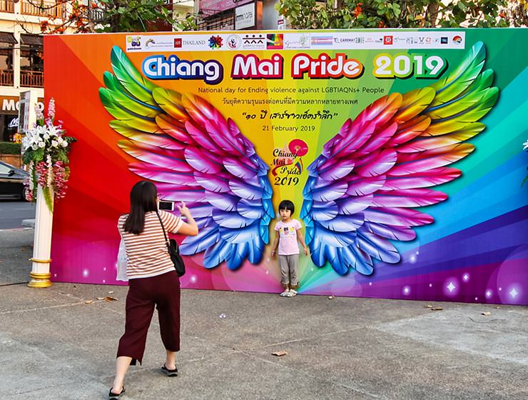 A mother taking a photograph of her child standing in front of the image of rainbow-colored wings at Chiang Mai Pride, gay parade in Chiang Mai, Thailand, photo by Ivan Kralj