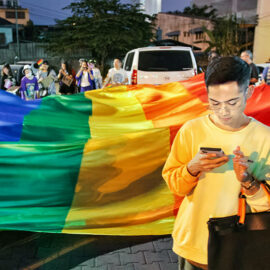 Young man looking at his mobile phone in front of the huge rainbow flag at Chiang Mai Pride, gay parade in Chiang Mai, Thailand, photo by Ivan Kralj