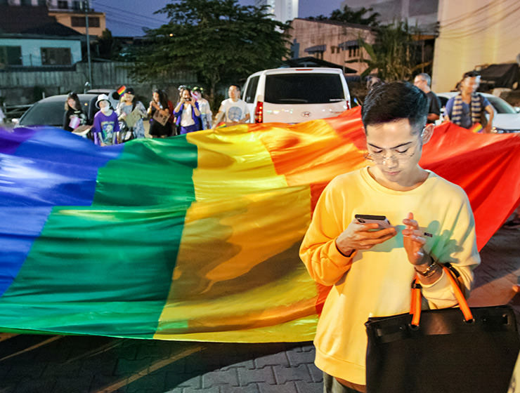 Young man looking at his mobile phone in front of the huge rainbow flag at Chiang Mai Pride, gay parade in Chiang Mai, Thailand, photo by Ivan Kralj