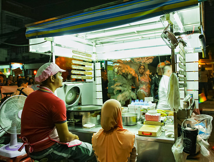 Participant with feathers at Chiang Mai Pride, gay parade in Chiang Mai, Thailand, passing by traditionally dressed roti makers' food station, photo by Ivan Kralj