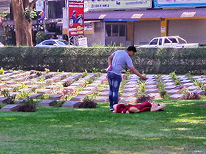 A woman laying in the grass at Kanchanaburi War Cemetery while a man is taking a photo of her from above, this inappropriate selfie was captured in Kanchanaburi, Thailand, photo by Ivan Kralj