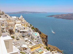 Whitewashed houses and swimming pools in Fira, the capital of Santorini, perched on the cliffs of caldera, with boats passing by, photo by Ivan Kralj