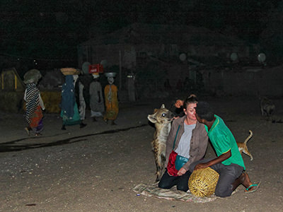 Hyena man feeding the hyena from the tourist's shoulder while local women disinterested pass by carrying goods on their hear, Harar, Ethiopia, photo by Ivan Kralj