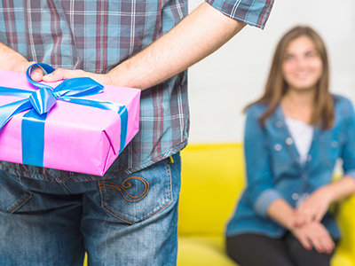 Man hiding a wrapped gift box behind his back, while a woman is waiting in the sofa, photo created by Freepik