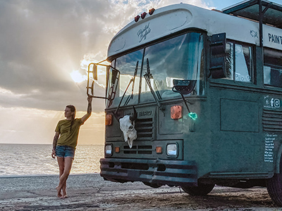 Jessica Rambo standing next to the Painted Buffalo Traveling Studio, a converted school bus, and her tiny house on wheels, with cloudy sky in the background
