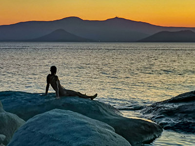 The silhouette of a naked man enjoying the sunset on the rock of Agia Anna Beach in Naxos, one of the best nude beaches in Cyclades Islands, Greece, photo by Nikos Samartzis