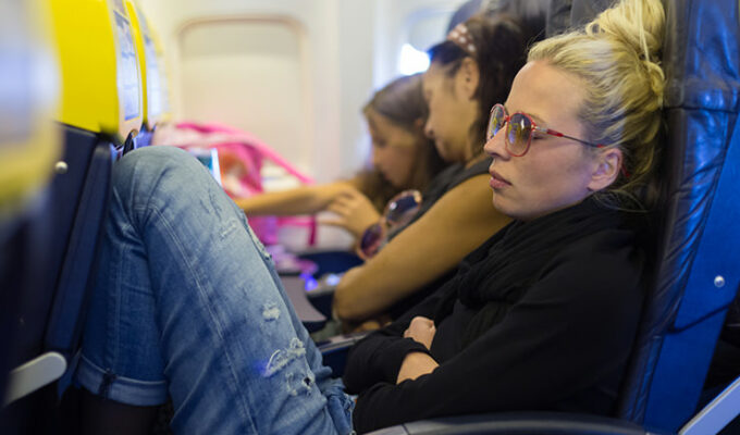 Tired woman napping on plane with legs up the seat, missing extra space. Modern planes offer less and less personal space to passengers, pushing them to purchase extra legroom seats. Photo by Kasto / Depositphotos