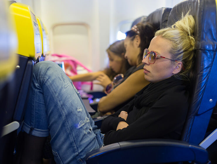 Tired woman napping on plane with legs up the seat, missing extra space. Modern planes offer less and less personal space to passengers, pushing them to purchase extra legroom seats. Photo by Kasto / Depositphotos