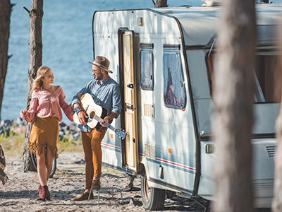 Hippie couple, a young woman dancing while a young man is playing the guitar next to the trailer parked in nature, photo by Artur Verkhovetskiy / Depositphotos.com