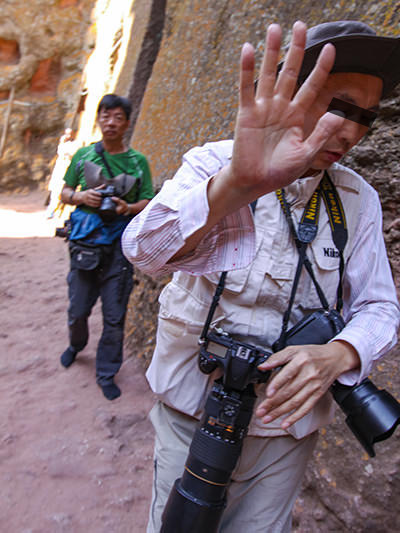 A Chinese photographer hiding behind his hand, after he was obsessively taking photographs of churchgoers at Bete Giyorgis church in Lalibela, Ethiopia, and then caught on camera by Pipeaway blogger Ivan Kralj who decided to "fight fire with fire", and turned the lens toward the perpetrator