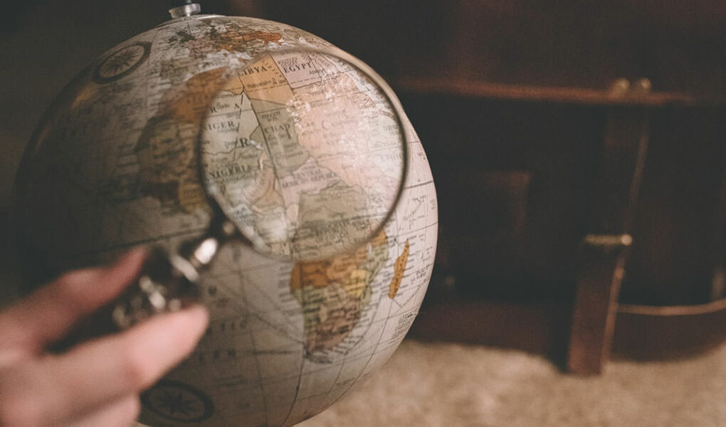 Globe model under magnifier with a suitcase in the background, photo by Clay Banks, Unsplash.com.