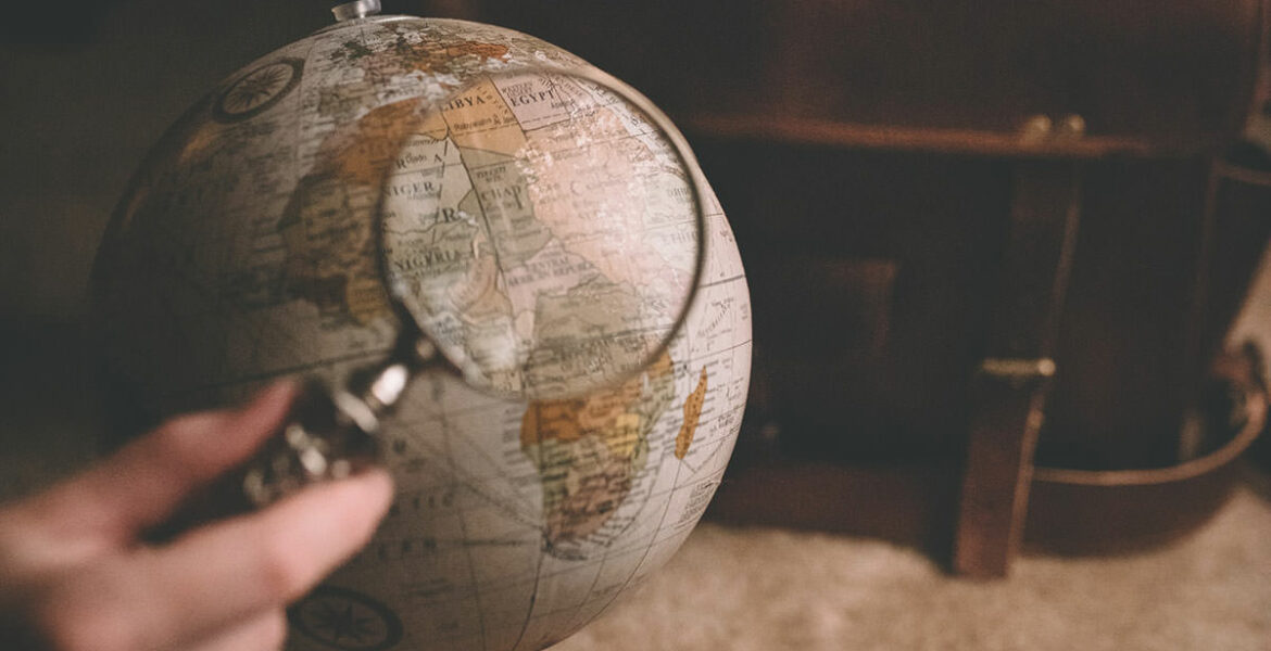 Globe model under magnifier with a suitcase in the background, photo by Clay Banks, Unsplash.com.