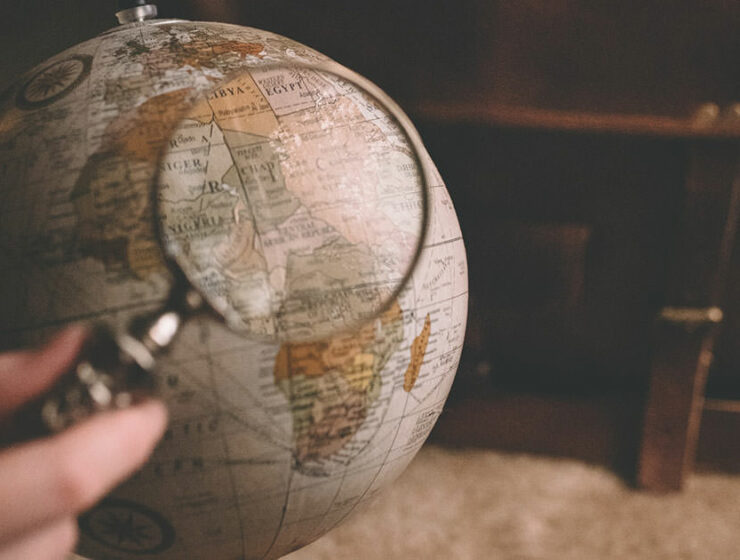 Globe model under magnifier with a suitcase in the background, photo by Clay Banks, Unsplash.com.