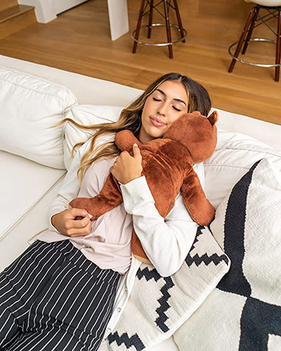 Young woman lying on a sofa with Darby the teddy bear on her chest, one of Hugimals brand of weighted stuffed animals.