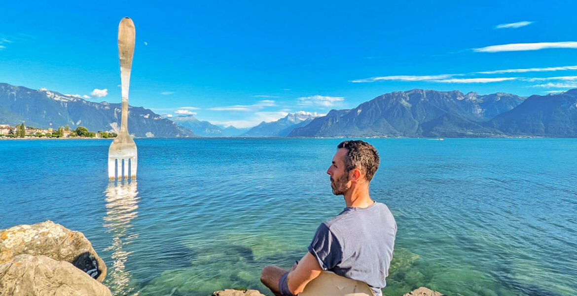 Travel blogger Ivan Kralj sitting on the bank of Lake Geneva, in Vevey, with the Fork of Vevey, 8-meter-tall steel fork in the lake, artwork by Jean-Pierre Zaugg, photo by Mladen Koncar.
