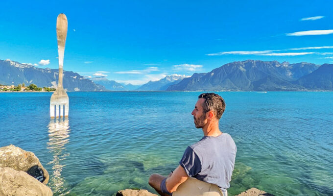 Travel blogger Ivan Kralj sitting on the bank of Lake Geneva, in Vevey, with the Fork of Vevey, 8-meter-tall steel fork in the lake, artwork by Jean-Pierre Zaugg, photo by Mladen Koncar.