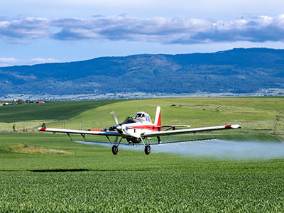 Ag pilot operating a crop duster plane above the field, spraying it with pesticides, photo by Eric Brehm, Unsplash.