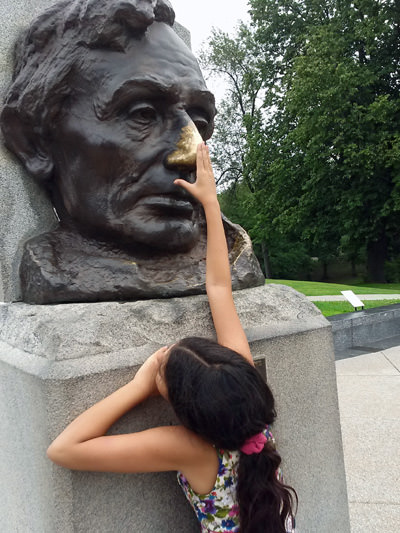 A girl reaching out with her hand to touch the "golden" nose of Abraham Lincoln's bronze statue that changed color due to excessive rubbing tradition for good luck, photo by Edenpictures.