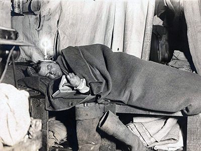 Artist George Marston, a member of the British Antarctic Expedition 1907-1909, lying in bed covered with a blanket, reading a book, and balancing a lighted candle on his head, copyright National Archives of Australia.