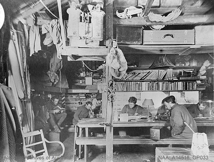 Men exploring Antarctica shot while reading and writing in Commonwealth Bay living hut during the Australian Antarctic Expedition 1911-1914, photo by Frank Hurley.