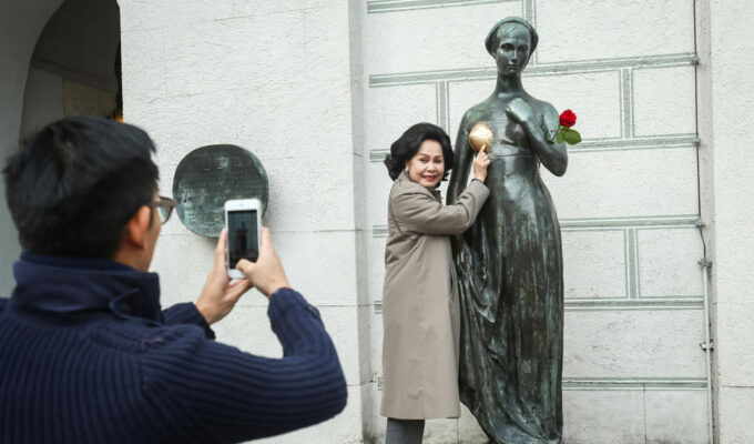 A woman posing for a photo standing next to the Juliet's statue in Munich, whose breast has changed color due to excessive statue rubbing by tourists, photo by Goran Jakus, Depositphotos.
