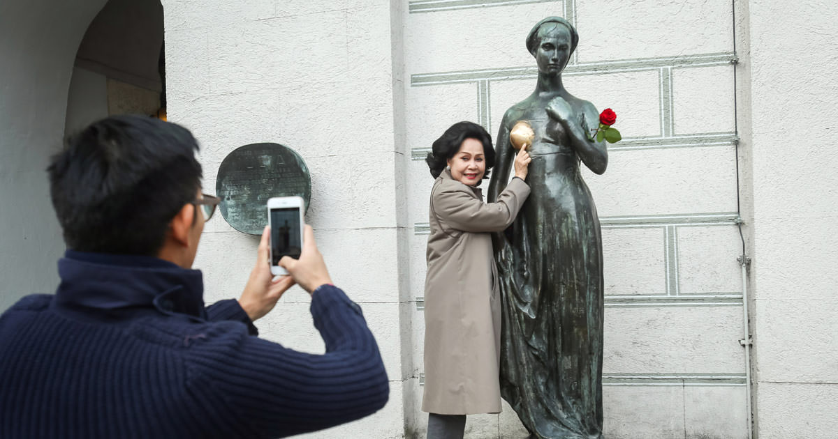 A woman posing for a photo standing next to the Juliet's statue in Munich, whose breast has changed color due to excessive statue rubbing by tourists, photo by Goran Jakus, Depositphotos.