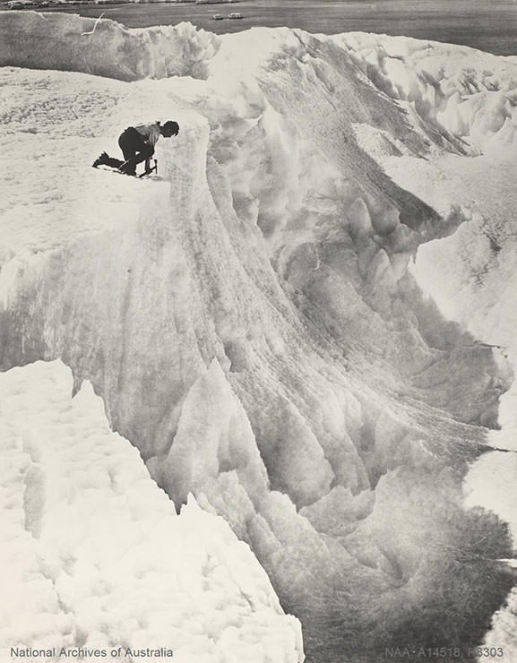 Man quarrying ice with a pick on the top of an ice cliff, shot during the Australian Antarctic Expedition 1911-1914.