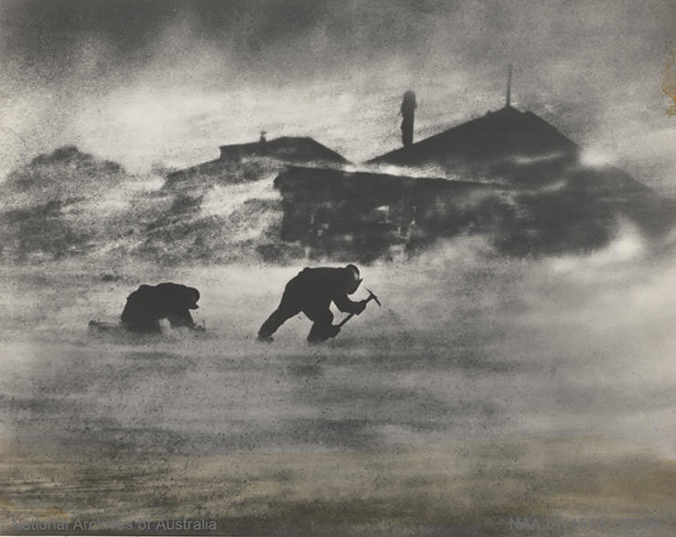 Two men quarrying ice with picks during a strong blizzard in front of their living huts in Antarctica, shot during the Australian Antarctic Expedition 1911-1914.