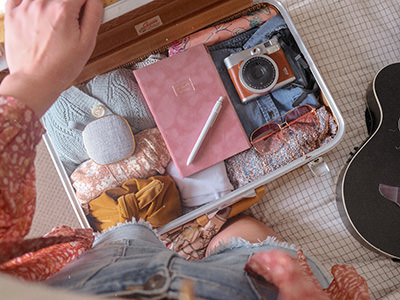 Packing a mini suitcase in pink color, and girly content, from light-colored clothes to a vitange camera, pink sunglasses and a journal; photo by Paige Cody, Unsplash.