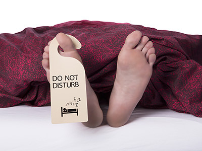 Feed poking out of the bed cover, with a DND sign hanging from the toe, an illustration of hotel 'Do Not Disturb policy violation; photo by joostverbeek, Depositphotos.