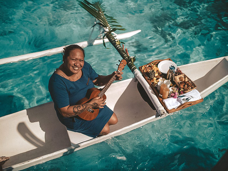 Woman playing a guitar while sitting in a canoe with breakfast; a special room service delivery from the sea at Intercontinental Bora Bora Resort, French Polynesia.