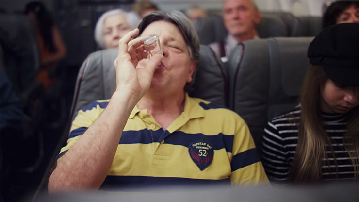 A plane passenger sitting in his seat and drinking alcohol from a small bottle, part of the Not On My Flight campaign against unruly passengers on flights; photo by EASA.