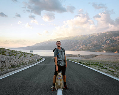 Tom Turcich and his dog Savannah standing in the middle of an empty island road in Croatia during their walk around the world.